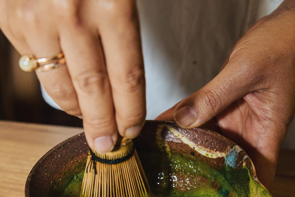 Hands using a bamboo chasen to whisk matcha in a ceramic bowl
