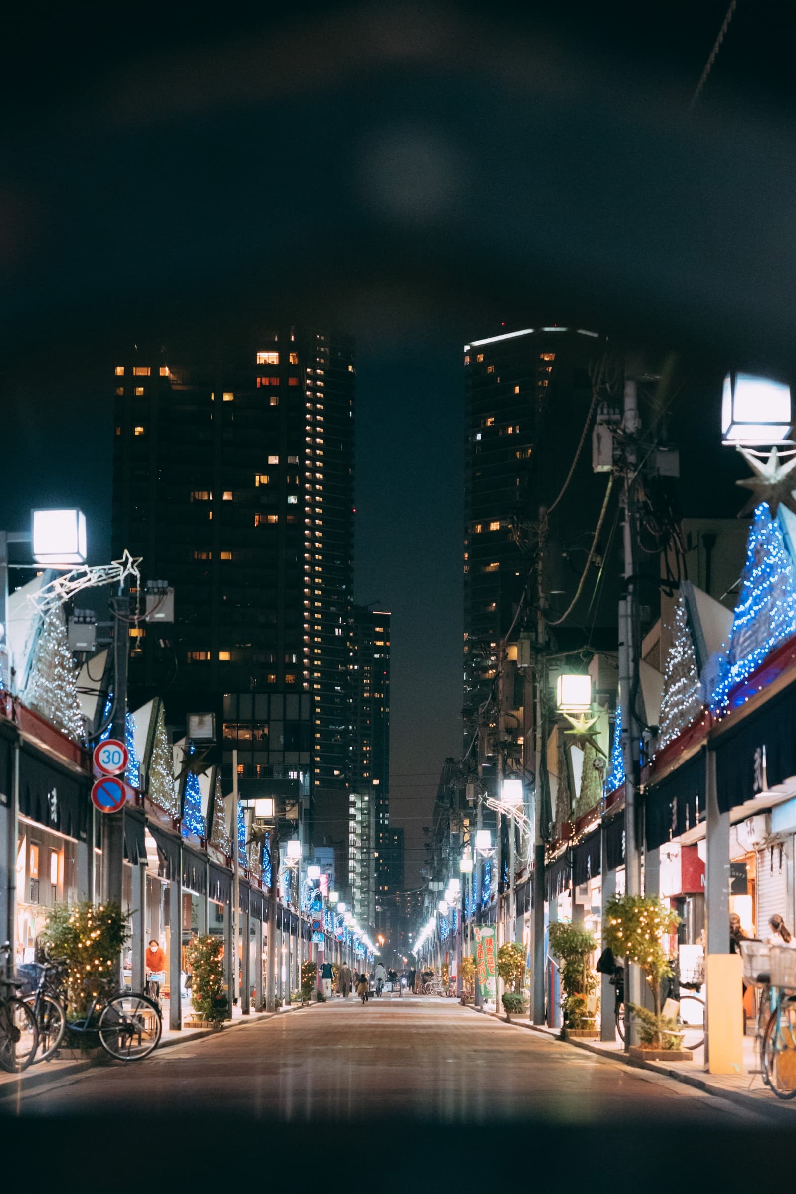 A symmetrical neighborhood shopping street at night in Tsukishima, Tokyo, with illuminated shop signs, parked bicycles, and residential towers visible behind the commercial frontage.