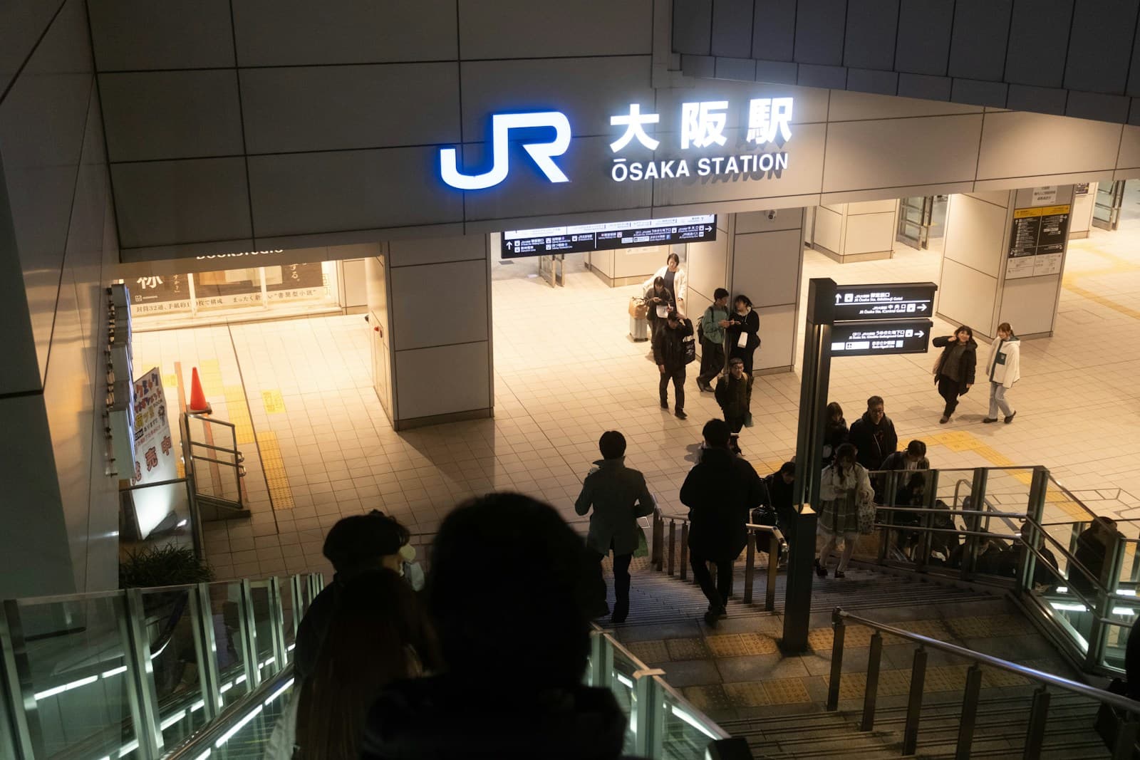 People enter and leave a brightly lit station entrance at night, with urban signage and street lighting visible.