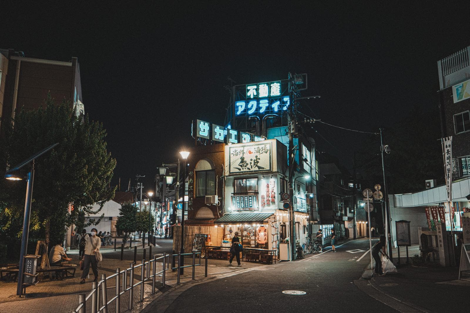 A retro neighborhood shopping street at night in Shiinamachi, Tokyo, with illuminated shop signs, storefront lights, and pedestrians on the sidewalk.