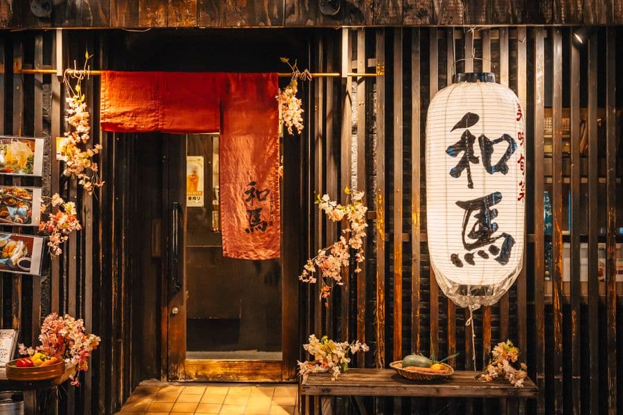 A traditional Japanese restaurant entrance at night with noren curtains, lanterns, and warm interior light spilling onto the sidewalk.
