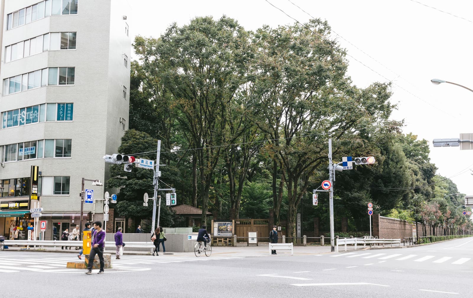An ordinary neighborhood intersection in Komagome, Tokyo, on an overcast early morning, with a cyclist crossing, a few pedestrians, shopfronts, and traffic signals before peak rush.