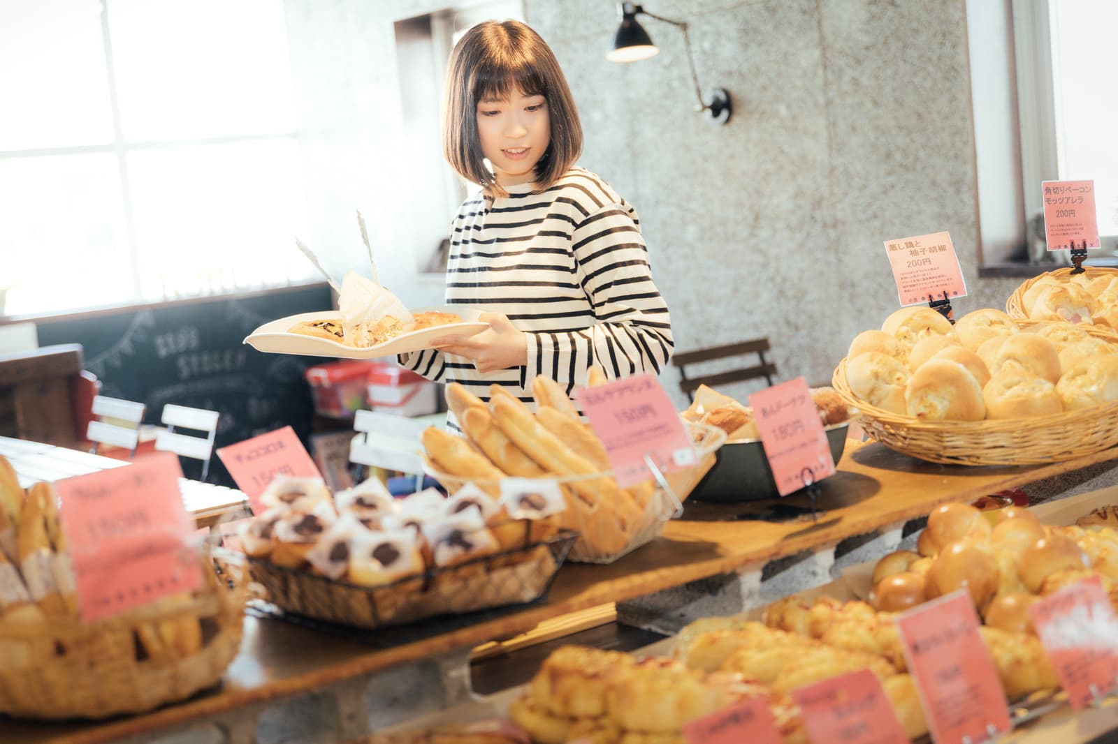 A customer holds a white tray and metal tongs while selecting round bread rolls from wooden display trays inside a Japanese bakery, with baguettes in a basket and a checkout counter visible behind.