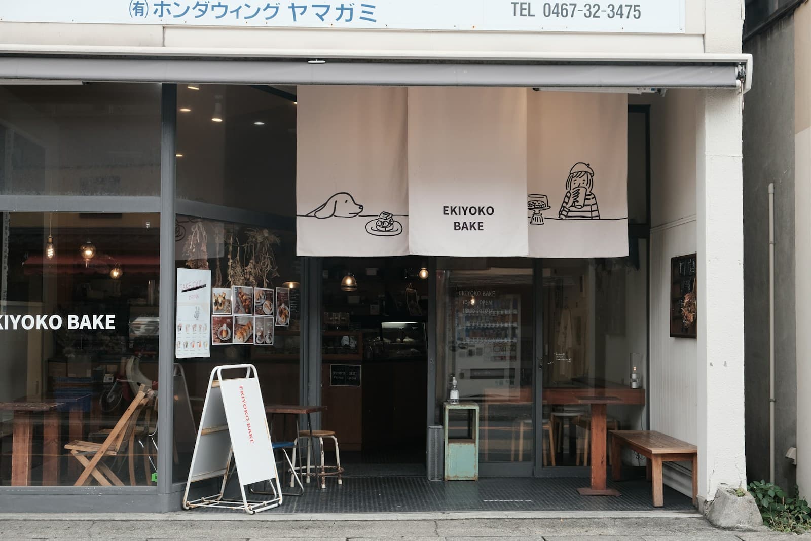 The EKIYOKO BAKE bakery storefront in Japan with white noren banners, pastry photos in the window, a take-out drink sign, and small wooden tables and a bench set on the sidewalk directly outside the entrance.