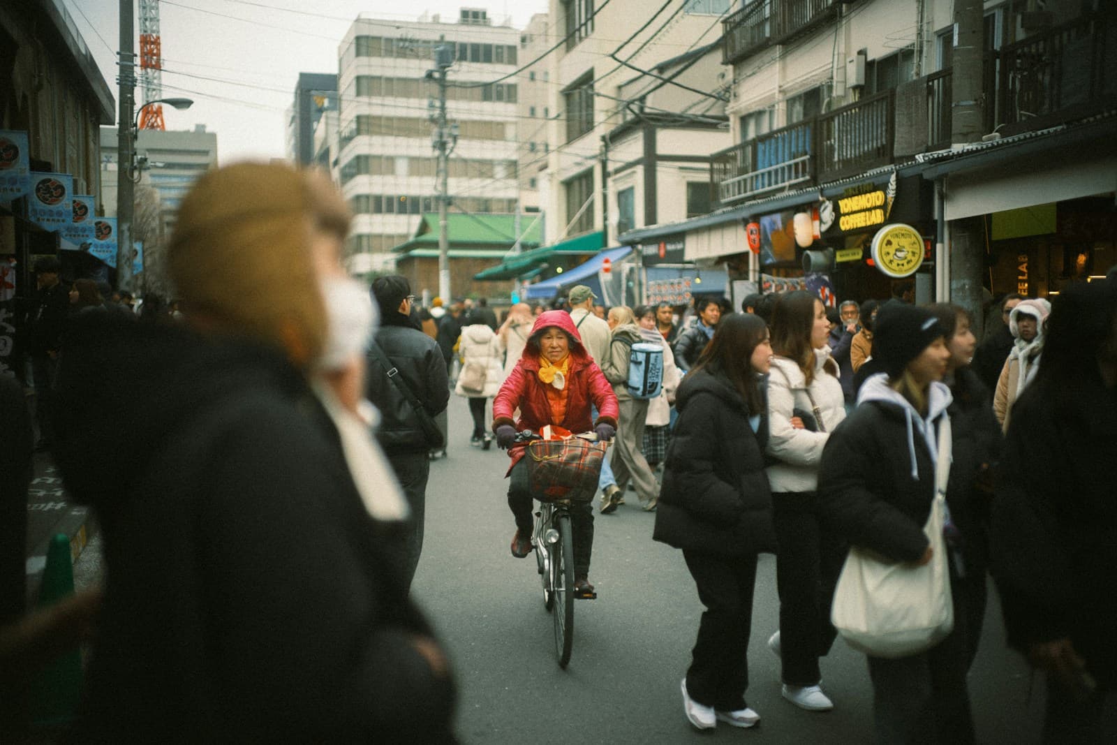 A cyclist rides through a busy Japanese shopping street in morning daylight, with pedestrians, shop signs, and storefronts along both sides.