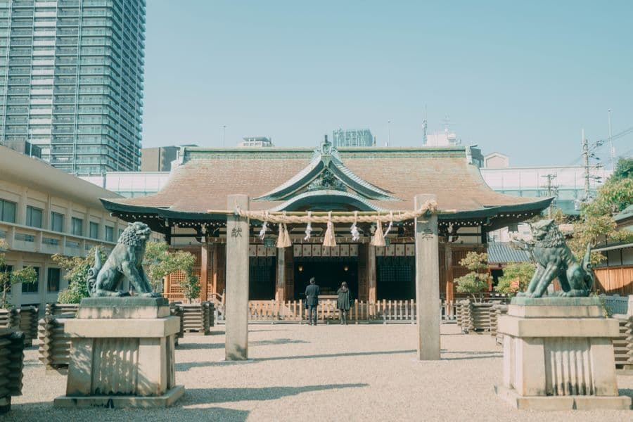 A traditional Japanese shrine set within an urban Tokyo neighborhood, with torii gate, stone steps, and mid-rise buildings visible beyond the sacred enclosure.