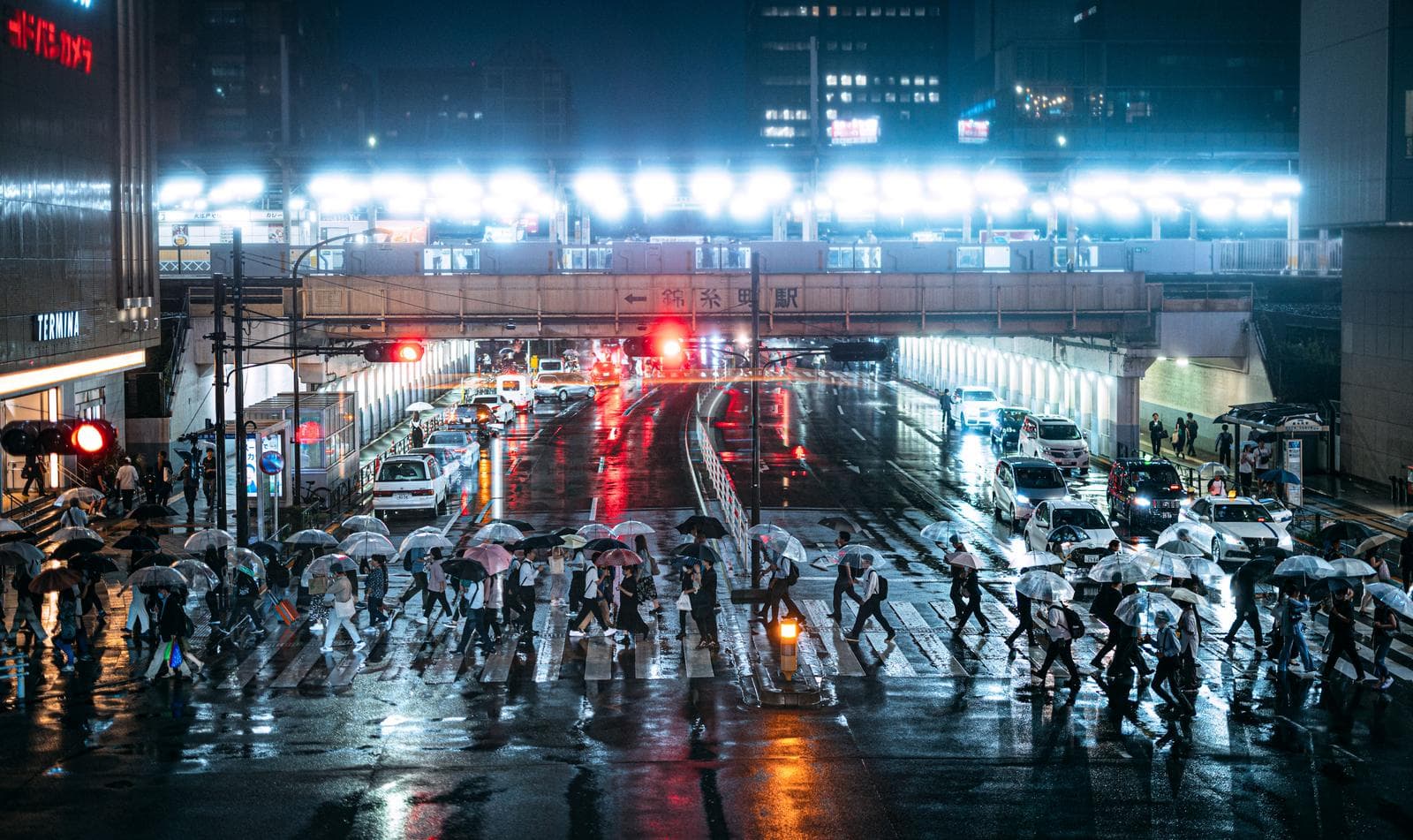 Pedestrians with umbrellas cross a rain-soaked intersection in Kinshicho, Tokyo, with wet pavement, traffic signals, and station-front buildings visible under active rainfall.