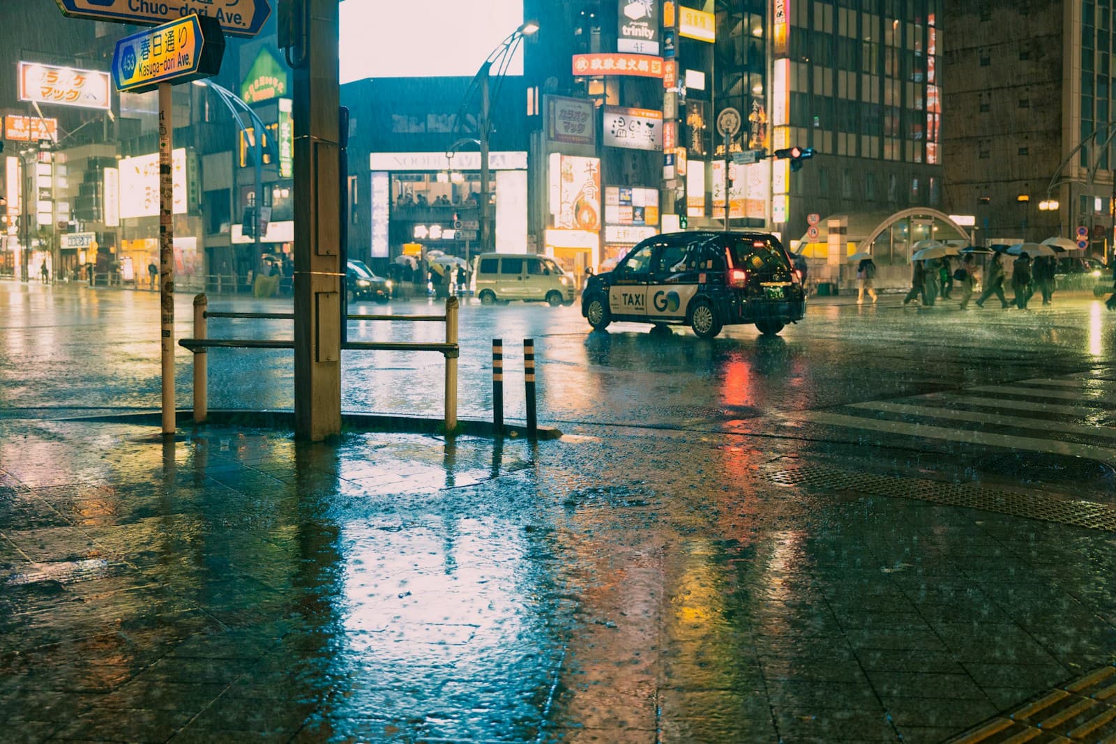 A wet urban intersection in the Ueno area of Tokyo during rain, with pedestrians holding umbrellas, a yellow taxi on reflective pavement, and street lights visible.