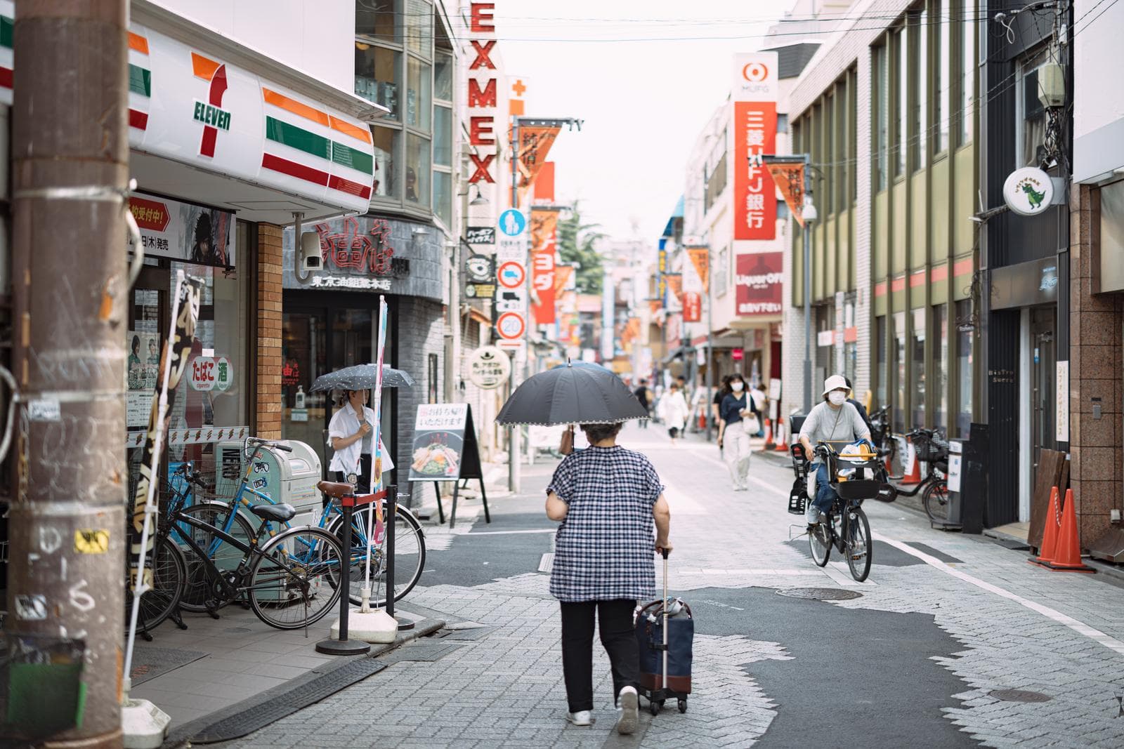 A covered neighborhood shopping street in Koenji, Tokyo, during rain, with a pedestrian holding an umbrella, wet pavement, and shop signs lining both sides.