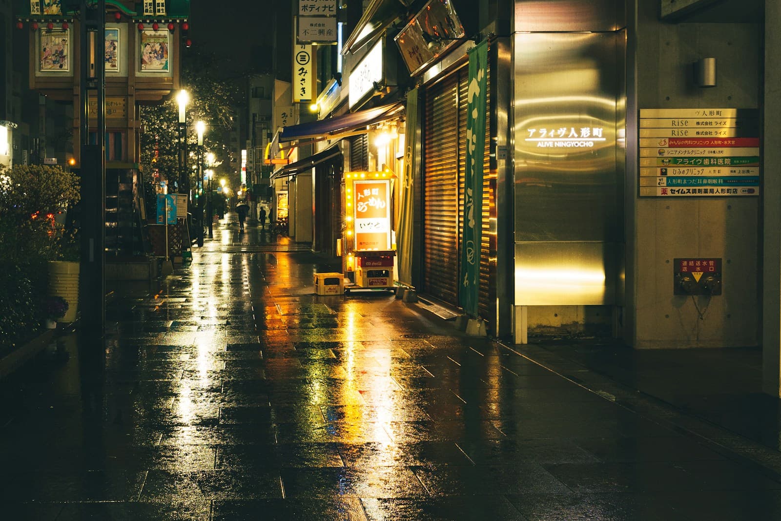 A wet commercial street at night in Tokyo during rain, with illuminated shop signs and their reflections visible on rain-darkened pavement.