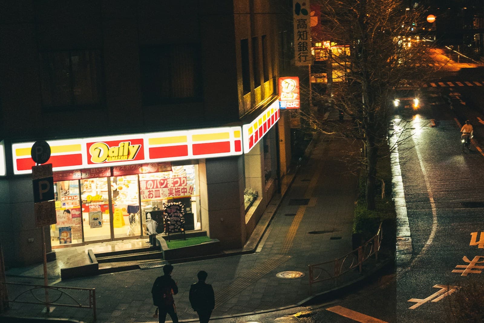Two pedestrians walk past a brightly lit convenience store on a wet Tokyo street at night, with rain-darkened pavement reflecting storefront light.