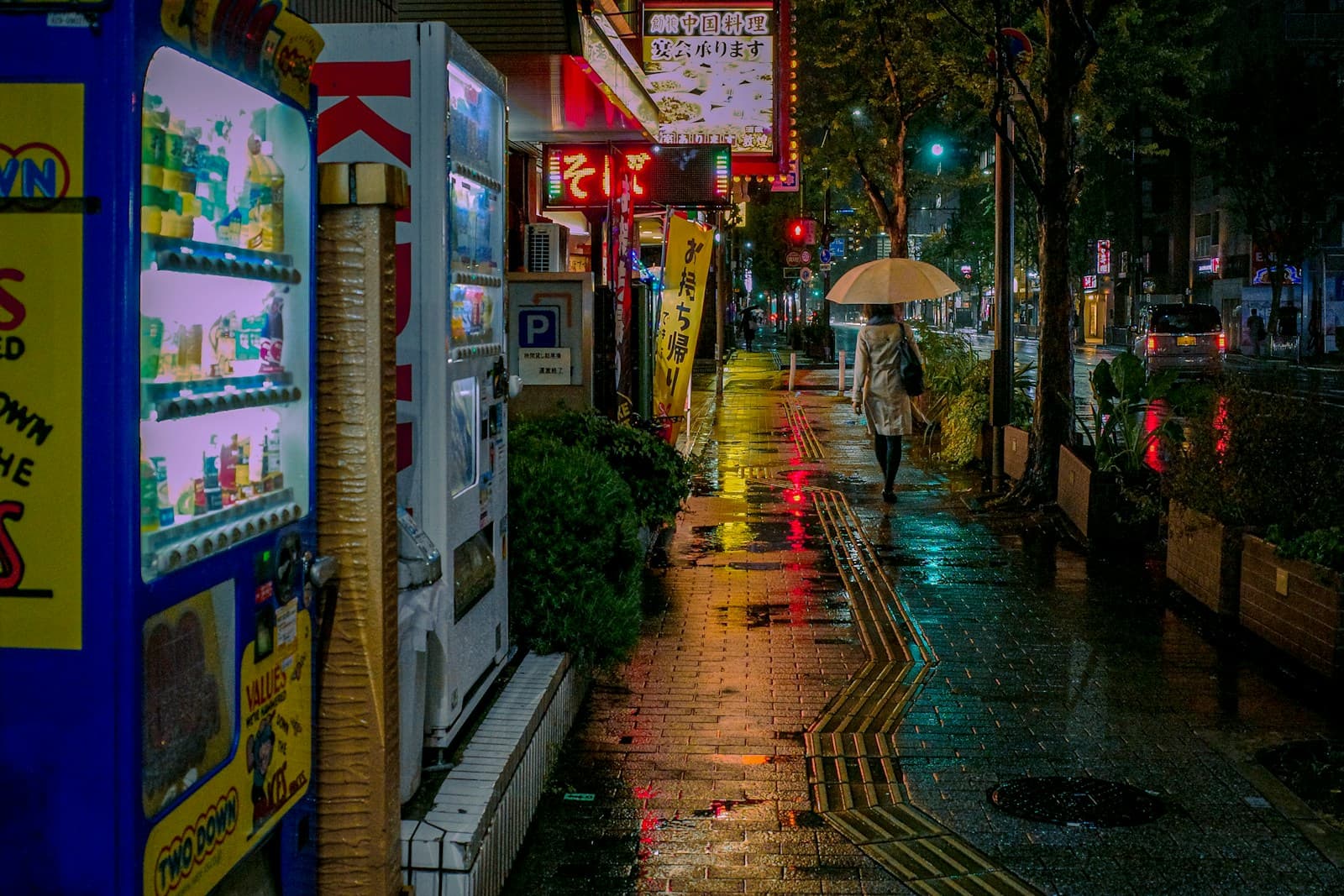 A person walks alone down a rain-soaked city street at night in Japan, holding an open umbrella with wet pavement reflecting street and shop light.