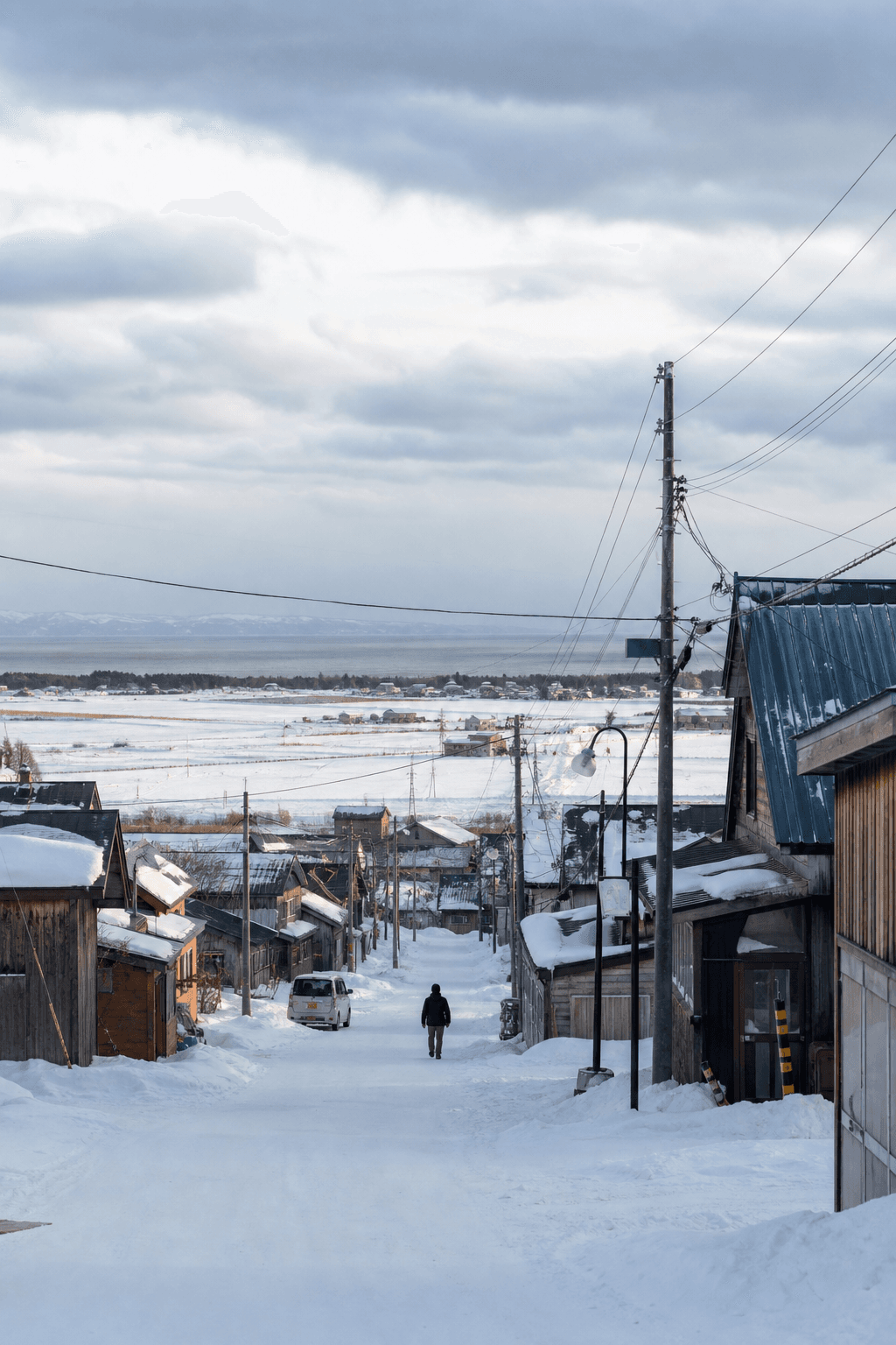 A quiet snowy street in a Hokkaido village with white fields and low wooden buildings under soft winter light