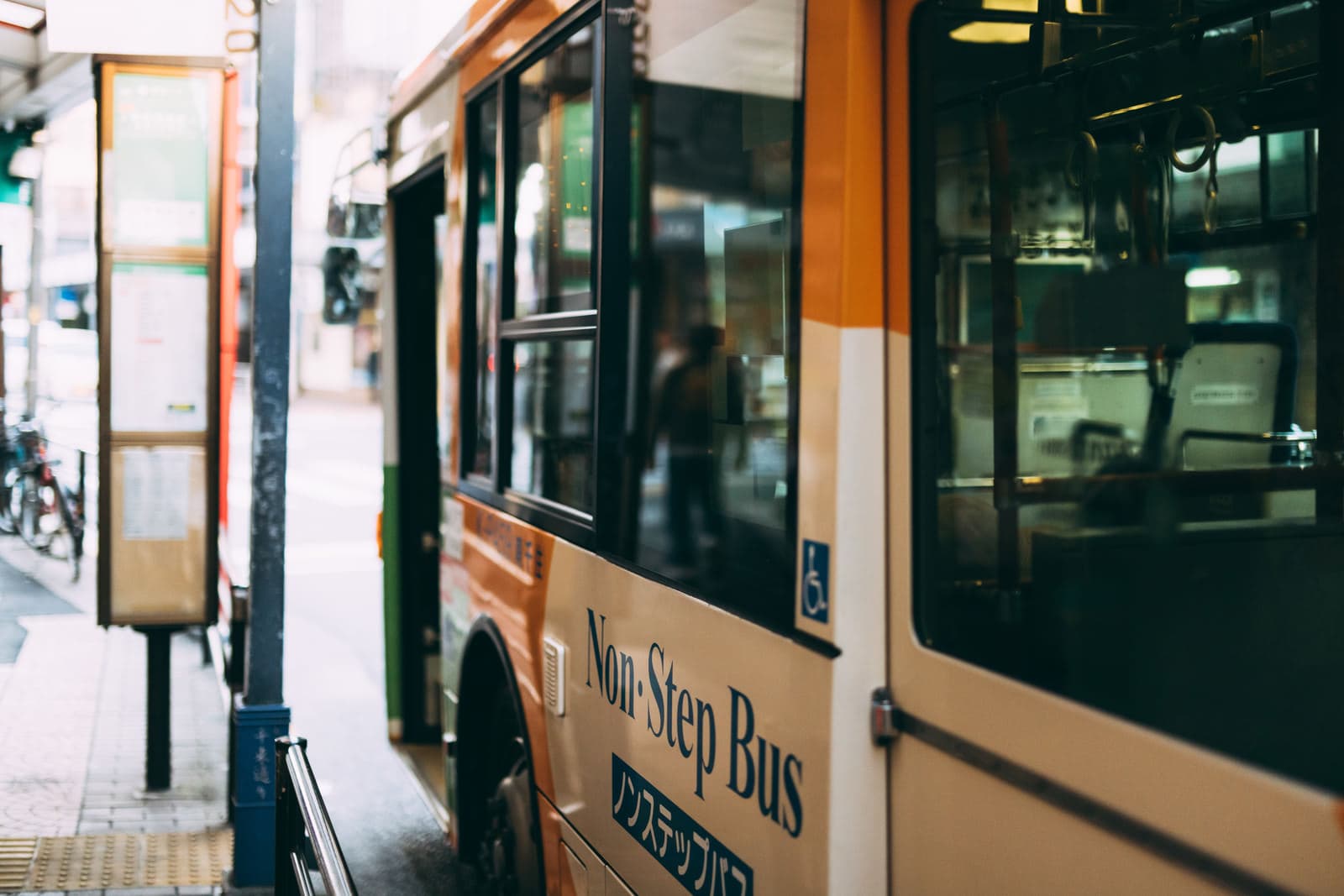 A green and beige non-step local bus stopped at a city bus stop with its door open for boarding