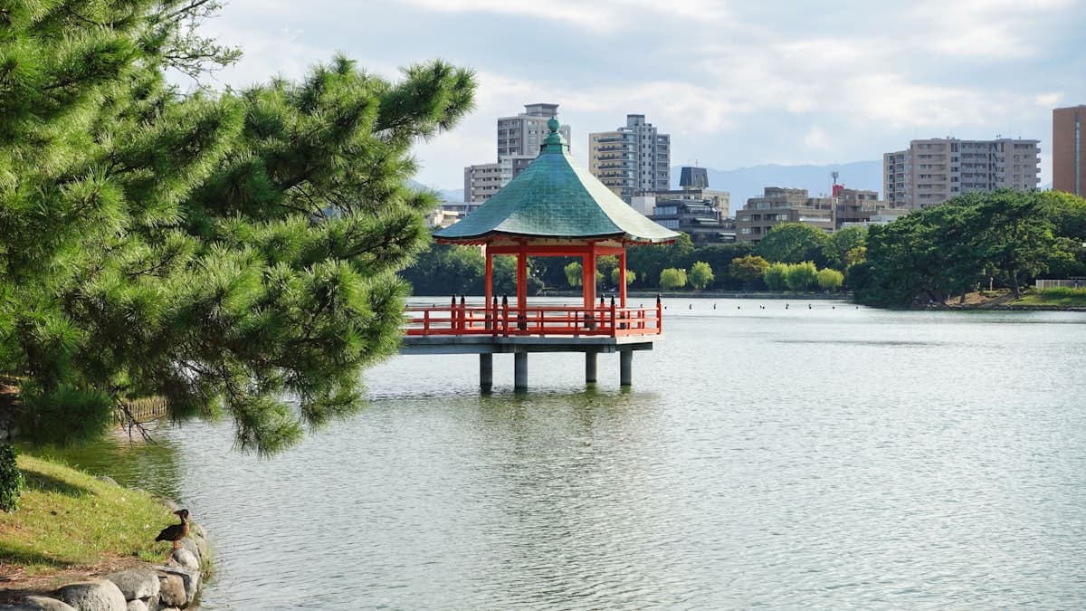 A traditional pavilion sits on Ohori Park pond with tree-lined shores and city buildings beyond.