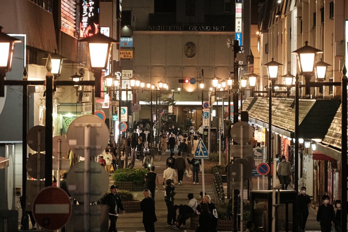 Pedestrians and vehicles move through a wide Tenjin intersection past commercial buildings under daylight.