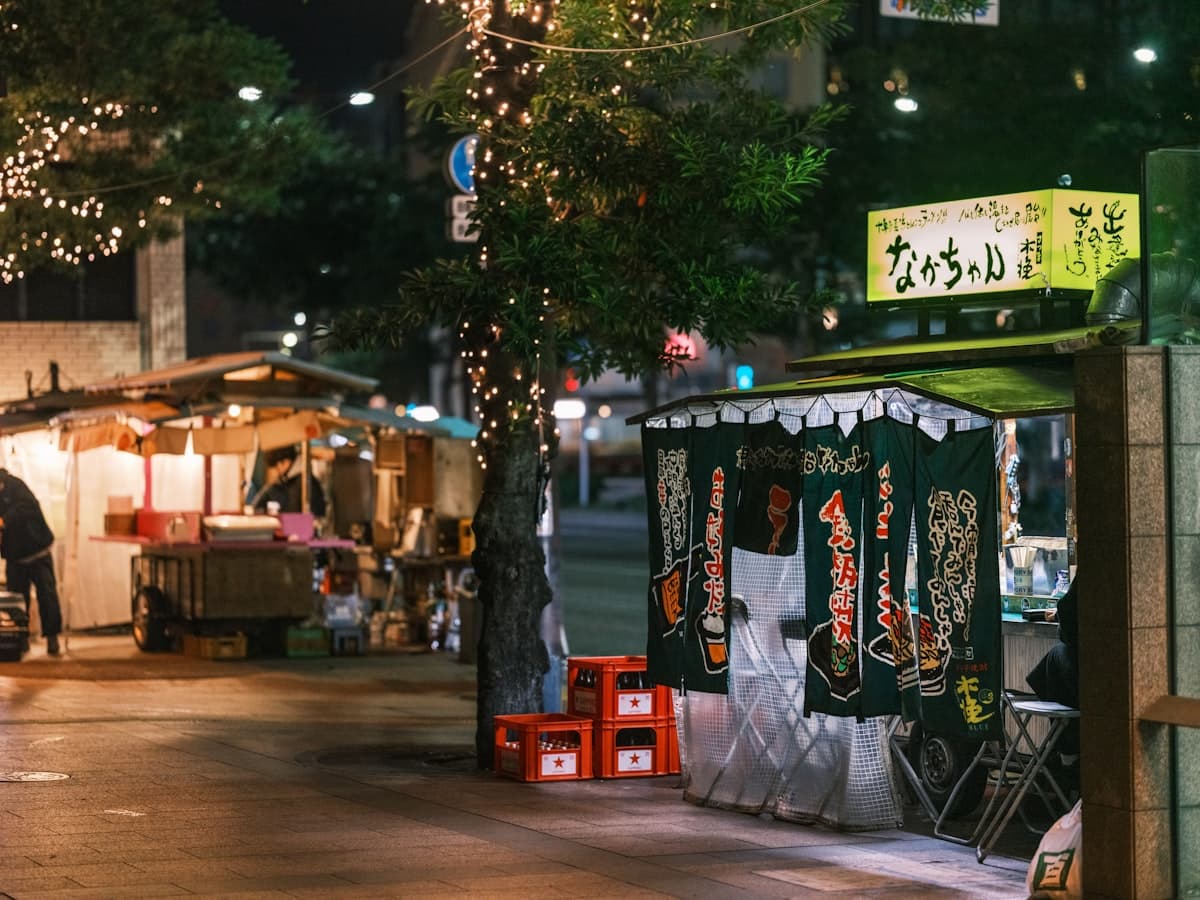 A traveler walks a Fukuoka street at night past lighted food-stall facades — an ordinary evening arrival moment, not a close-up of a single counter.