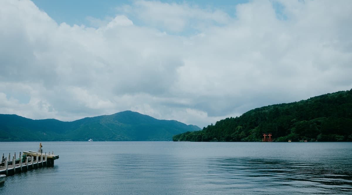 Lake Ashi stretches toward forested mountains under soft cloud cover, with a wooden pier in the foreground.