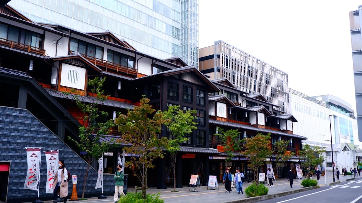 Pedestrians walk a wide sidewalk past traditional-style shopfronts and modern buildings in Hakone-Yumoto.