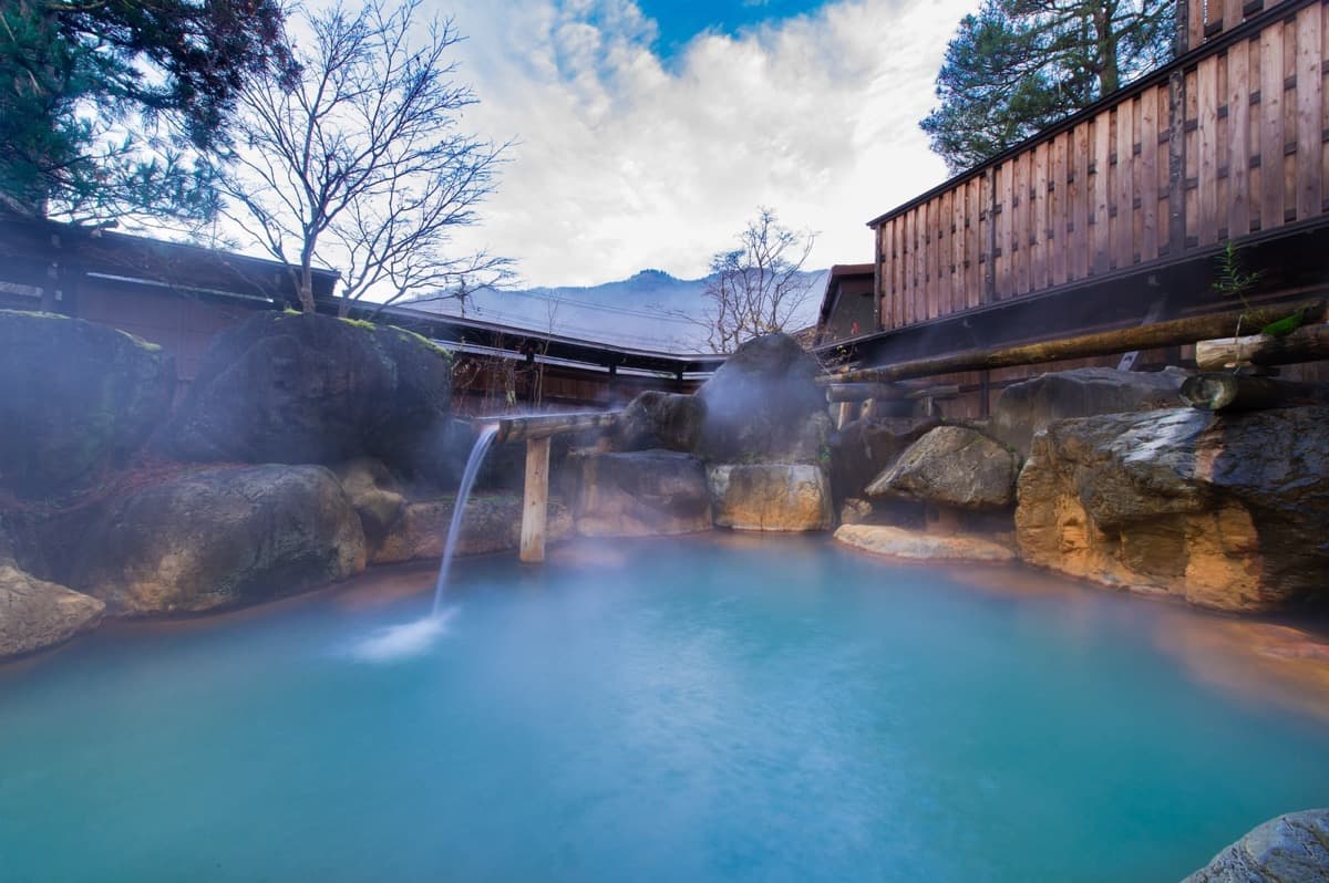 Steam rises from a rock-lined outdoor hot spring bath enclosed by wooden fences, with misty mountain slopes beyond — a quiet ryokan rotenburo moment at dusk or dawn.