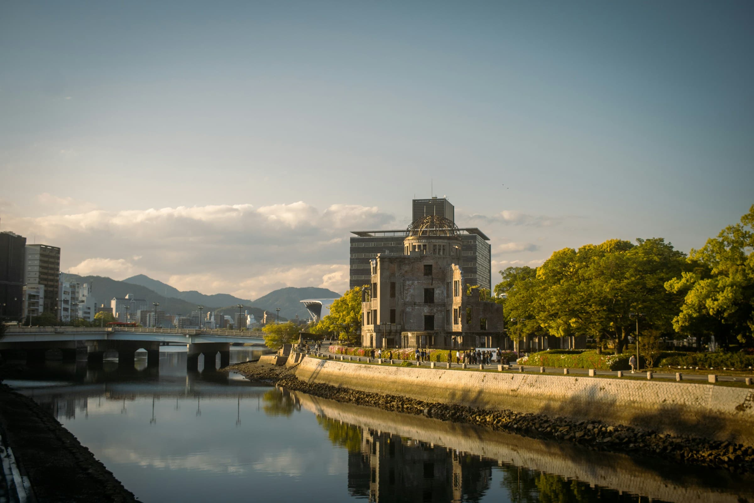 Hiroshima cityscape at golden hour, with the Atomic Bomb Dome and modern buildings along the Motoyasu River reflected in the water and mountains beyond the skyline.