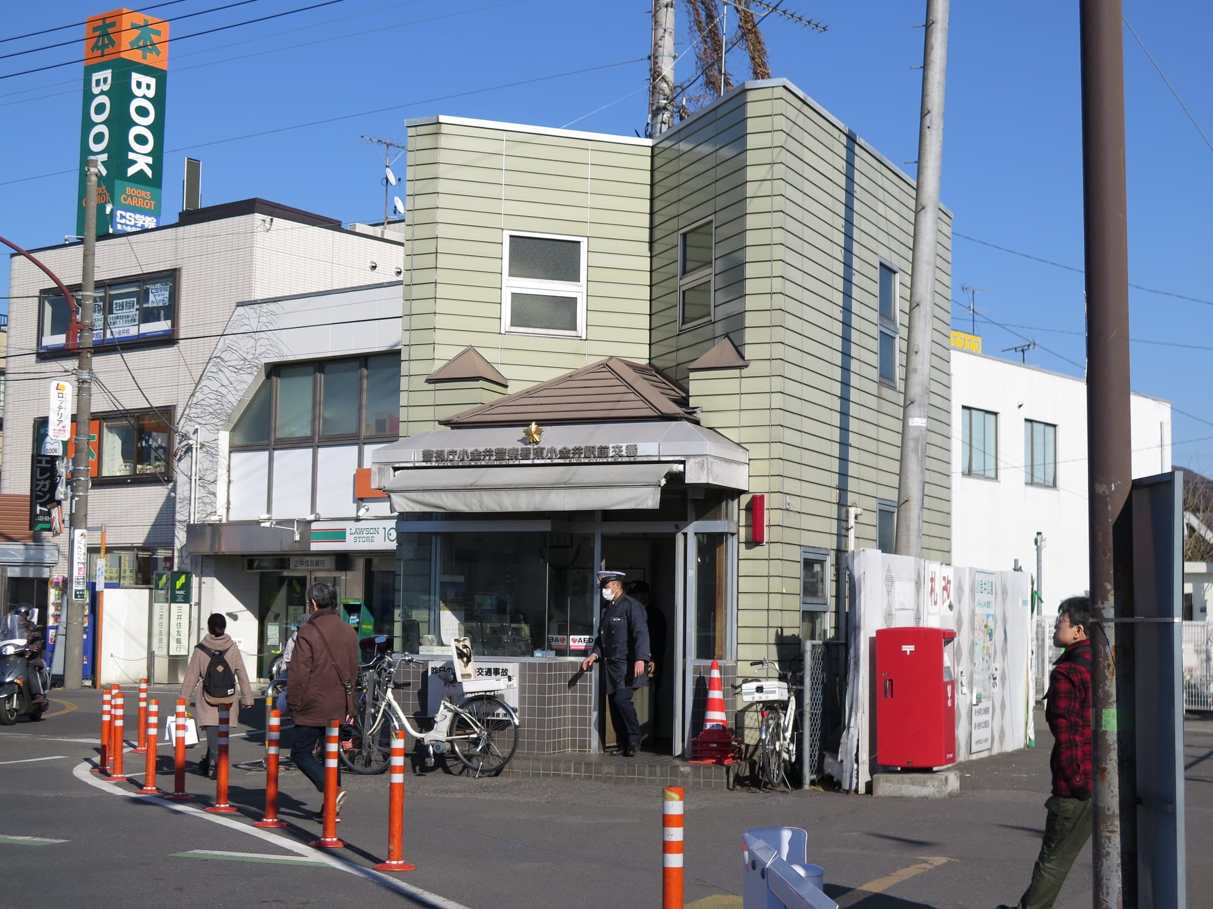A neighborhood police box (koban) with an officer standing at the entrance, on a daylight street corner near a station.