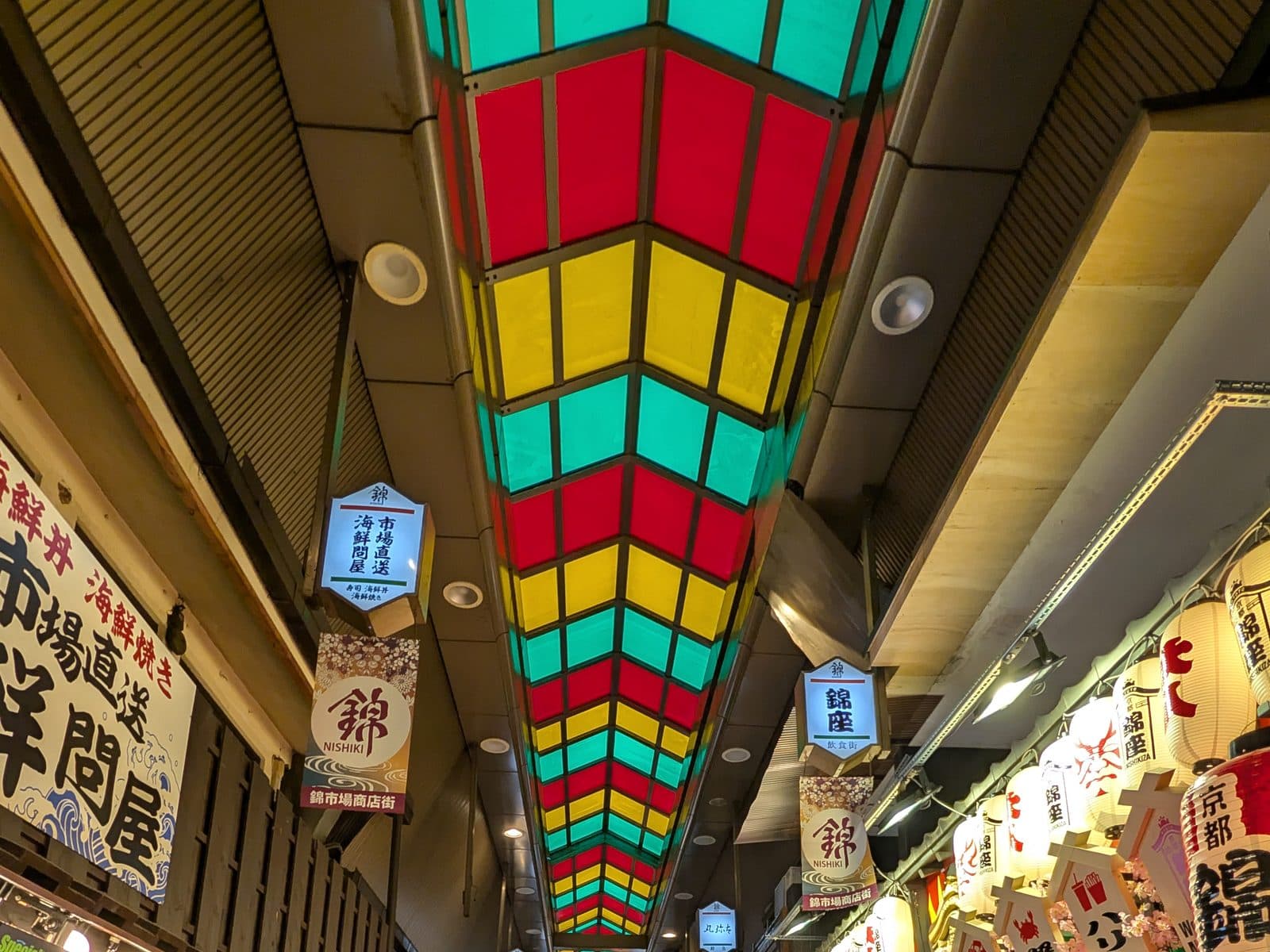 A colorful diamond-patterned ceiling canopy above the covered Nishiki Market arcade in Kyoto, with shop banners and lanterns lining both sides.