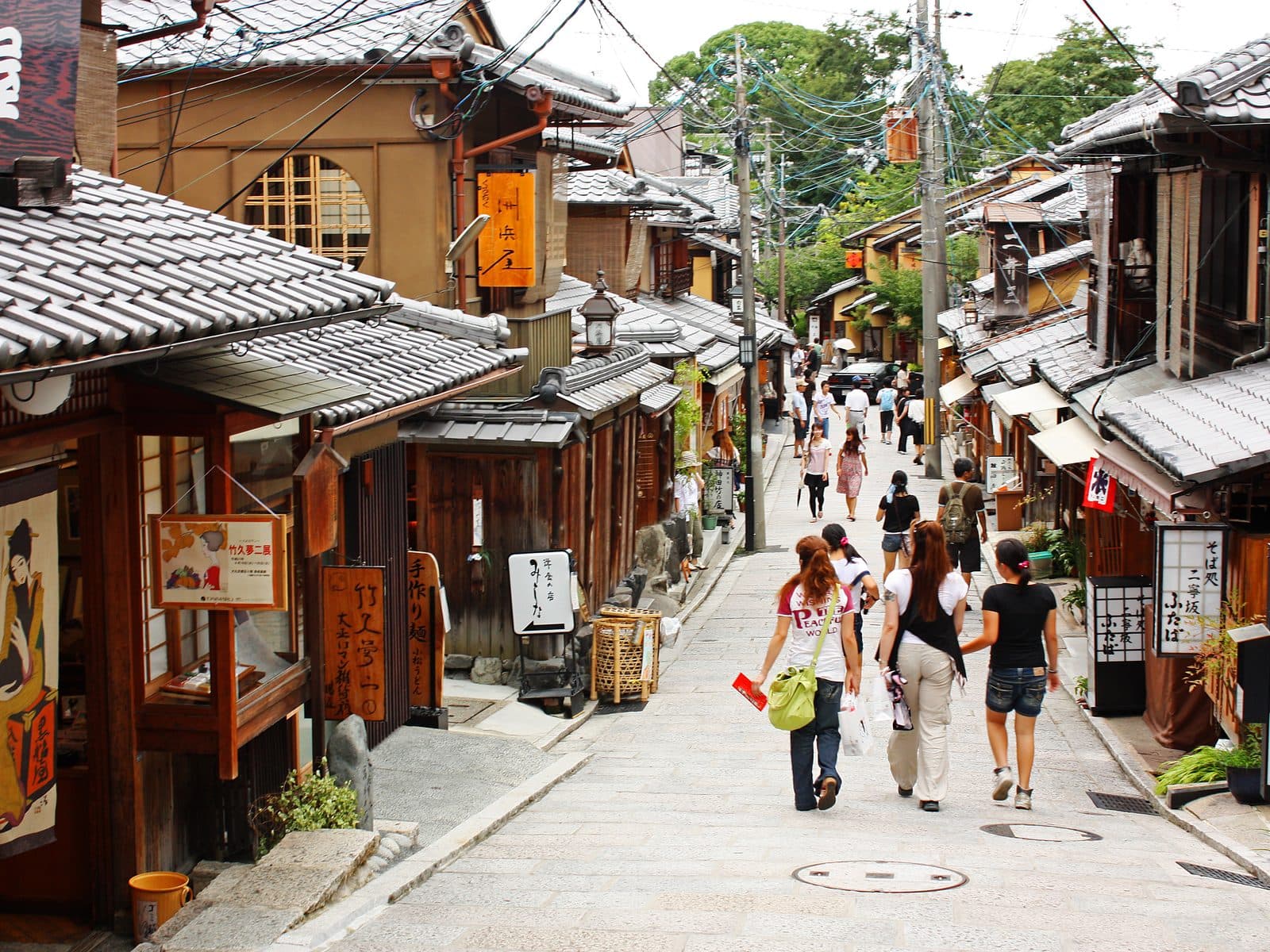 An elevated view down the sloped, stone-paved Sannenzaka street in Kyoto's Higashiyama district, lined with traditional wooden shopfronts and a few pedestrians.