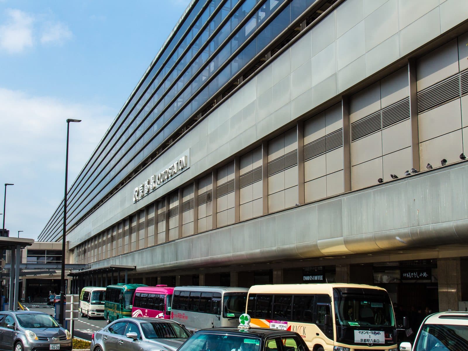 Kyoto Station's large glass-and-steel exterior facade with bilingual signage and tour buses parked in front, under a clear sky.