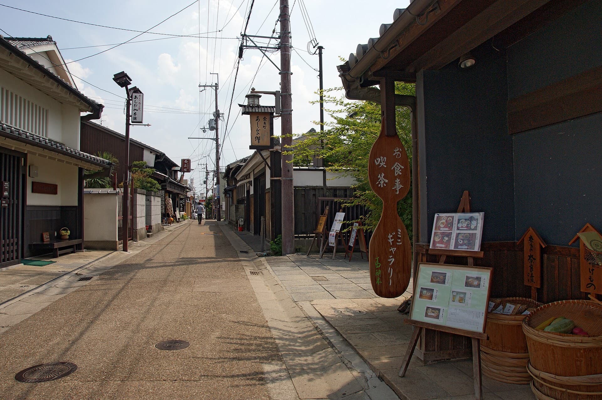 A quiet preserved street in Naramachi, Nara, lined with traditional wooden machiya buildings and shop signs, with a pedestrian walking in the distance.