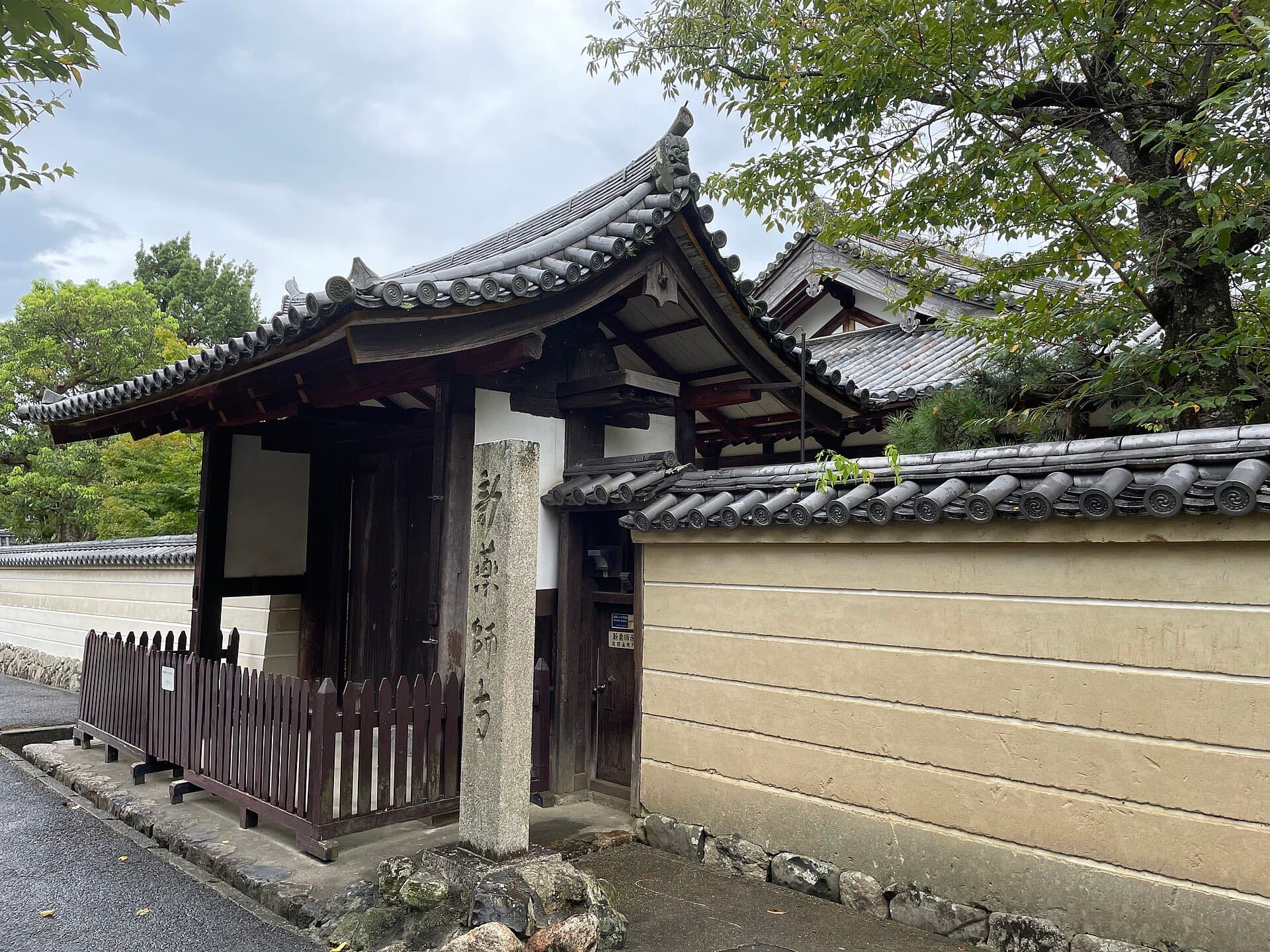 The quiet gate and stone marker of Shin-Yakushiji temple in the Takabatake district of Nara, surrounded by trees and a residential street.