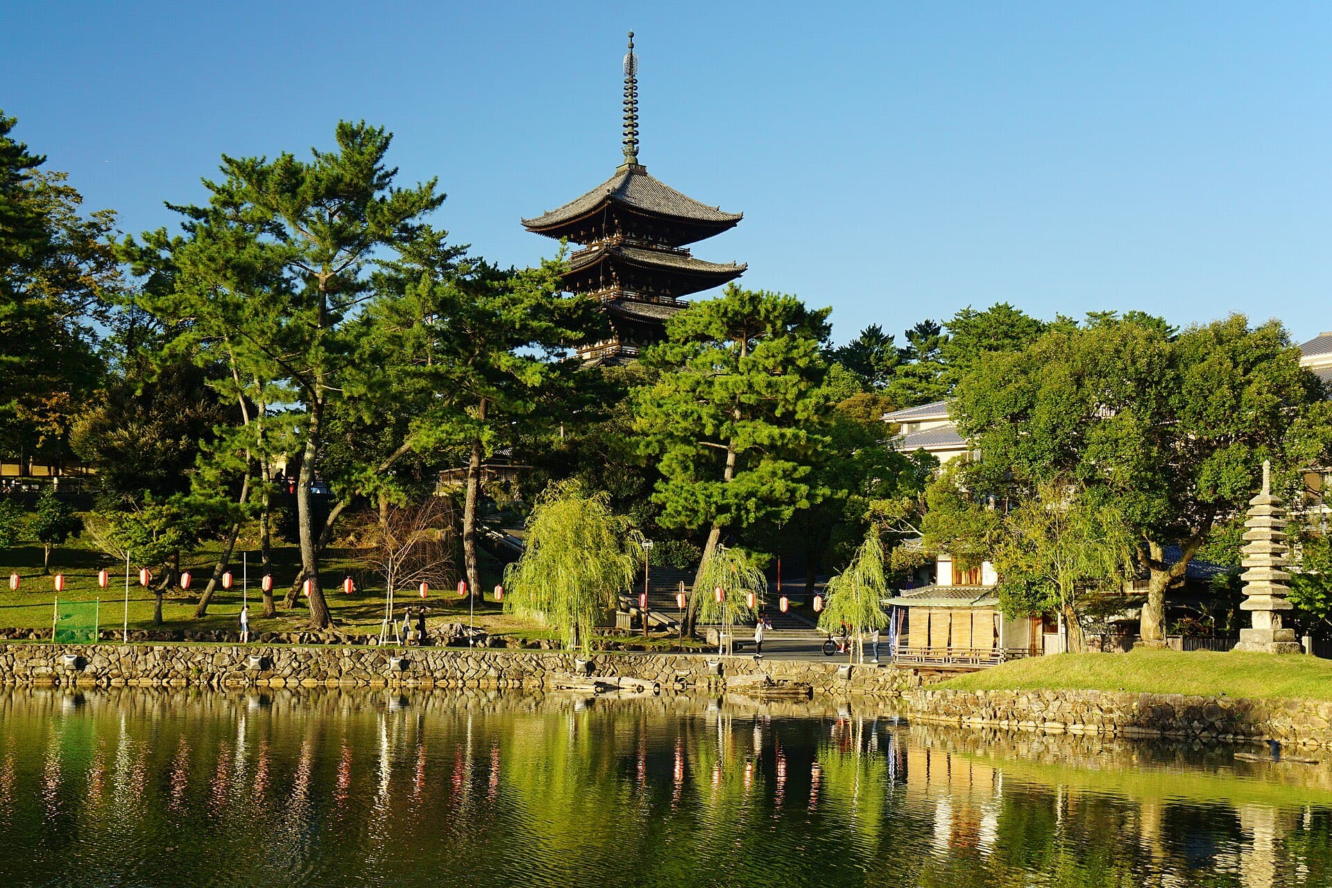The five-story pagoda of Kofuku-ji reflected in Sarusawa Pond in Nara, framed by pine and willow trees under a clear blue sky.