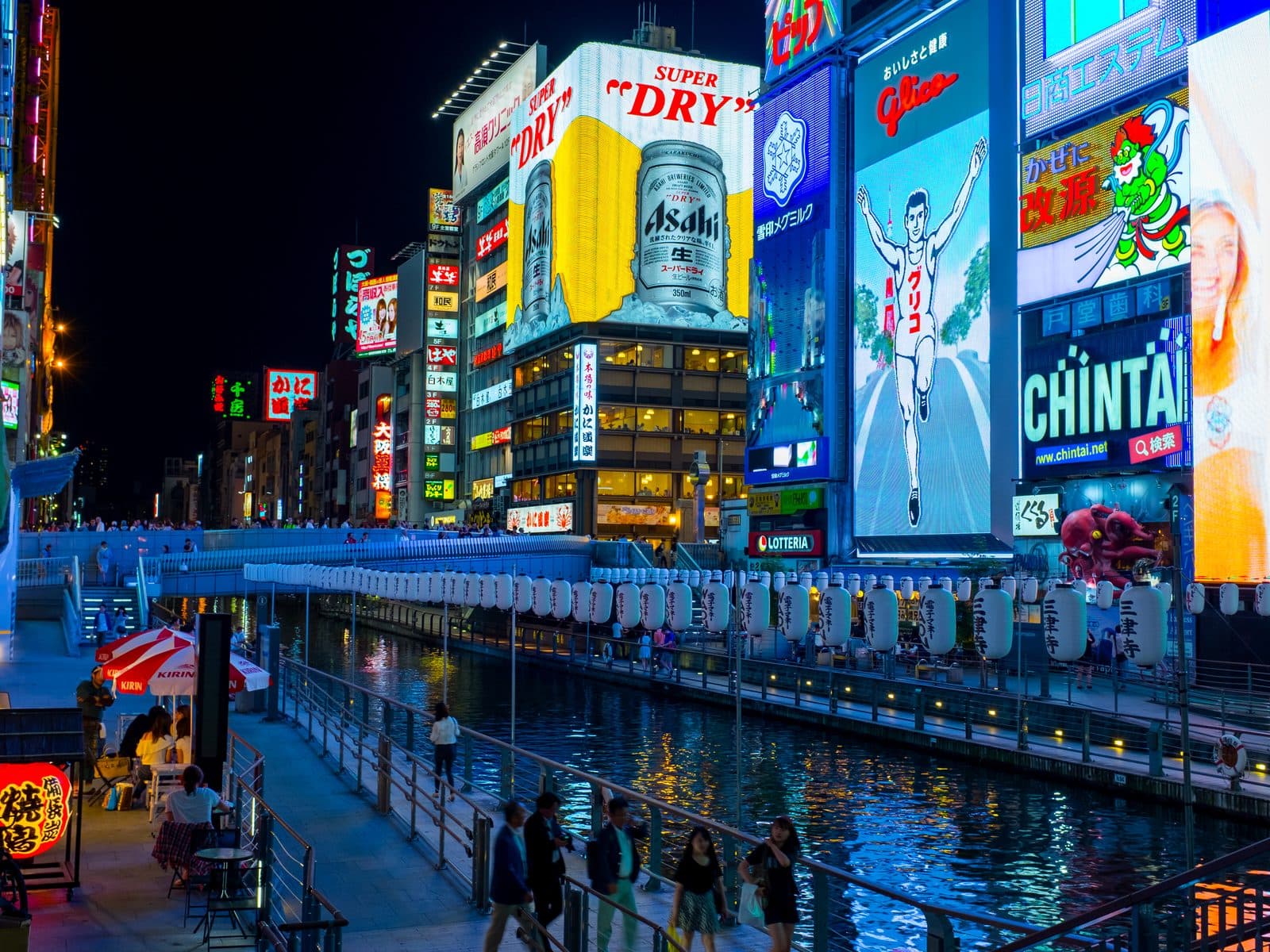 Dotonbori canal at night in Osaka, with the illuminated Glico running man sign, neon storefronts, and pedestrians along the canal-side walkway.