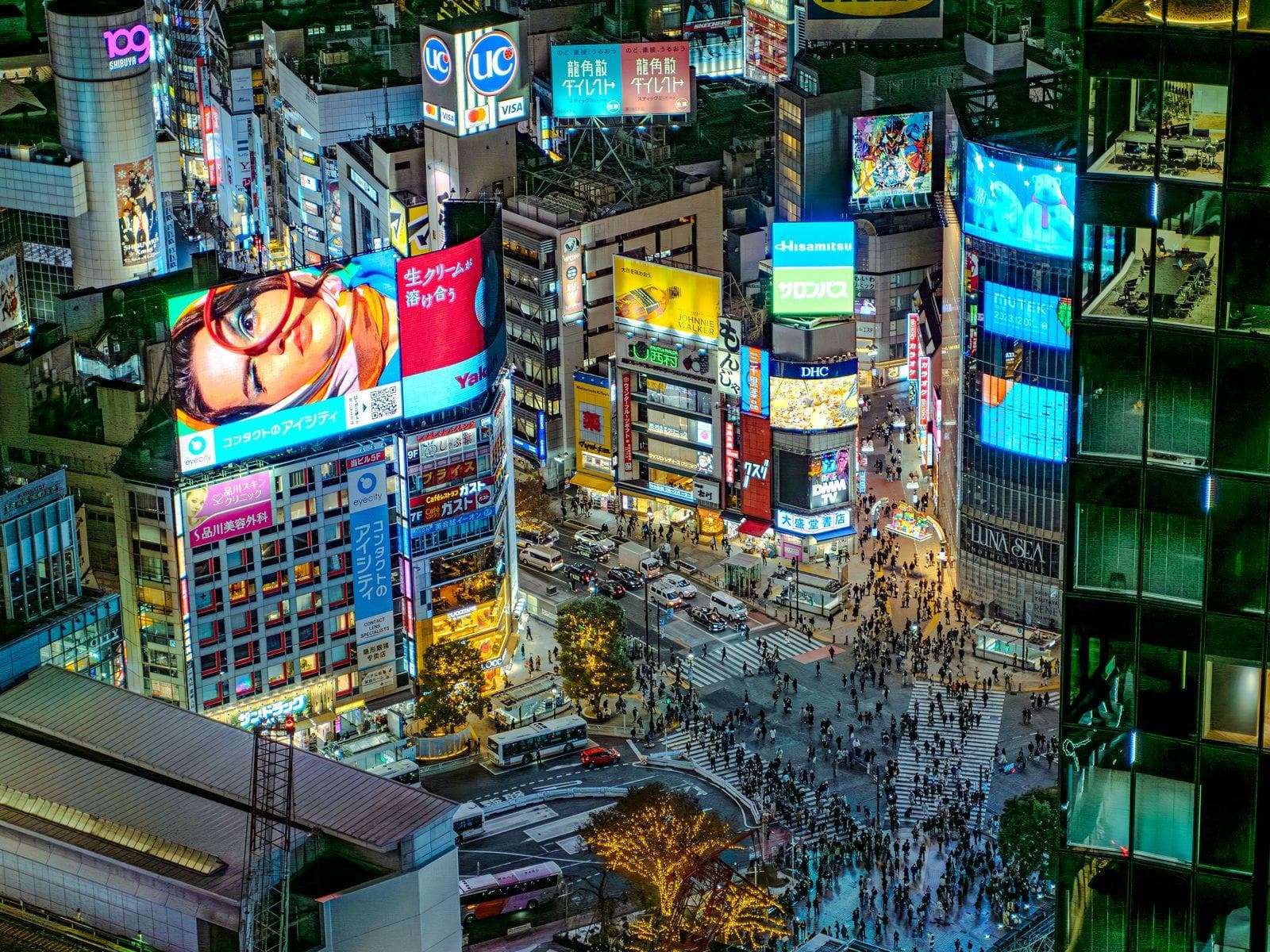 An elevated night view of Shibuya Scramble Crossing surrounded by illuminated buildings and digital advertising screens, with pedestrians crossing below.
