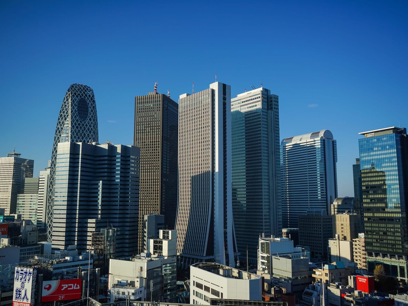 Shinjuku's skyscraper skyline in daylight, including the distinctive lattice-patterned Mode Gakuen Cocoon Tower, under a clear blue sky.