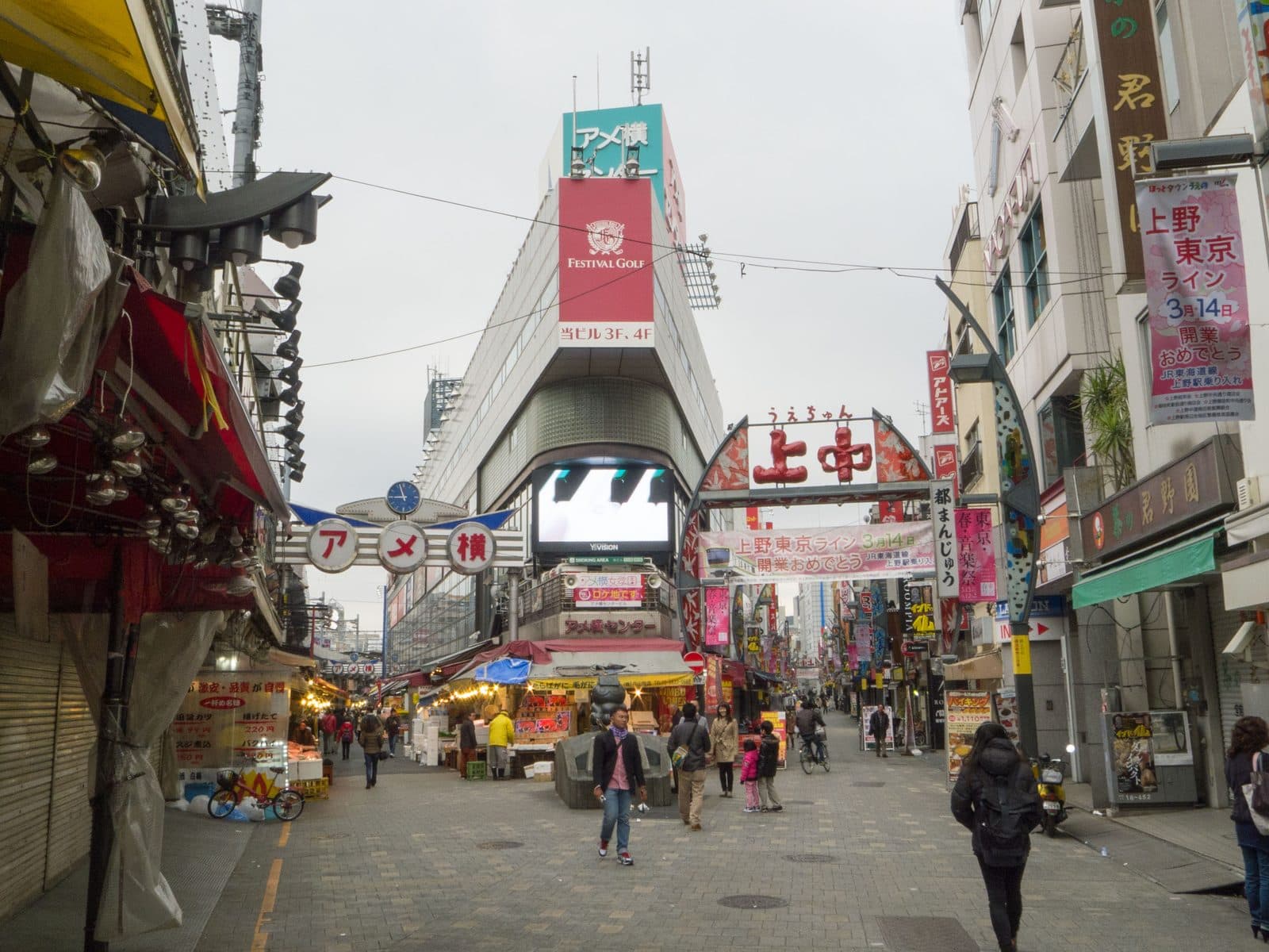The Ameyoko market street near Ueno Station, lined with shop awnings and signage beneath the overhead "アメ横" arch, with a few pedestrians walking through.