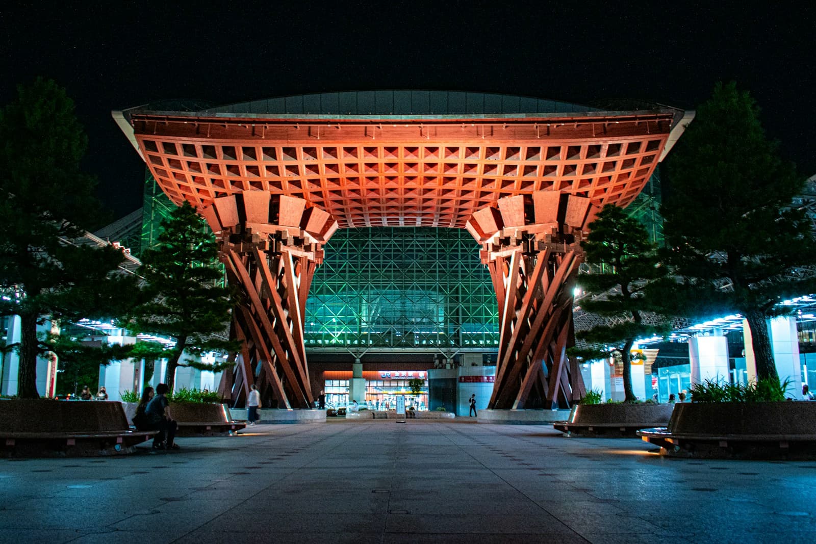 The wooden Tsuzumi-mon gate at Kanazawa Station at night, with trees framing the illuminated entrance structure.
