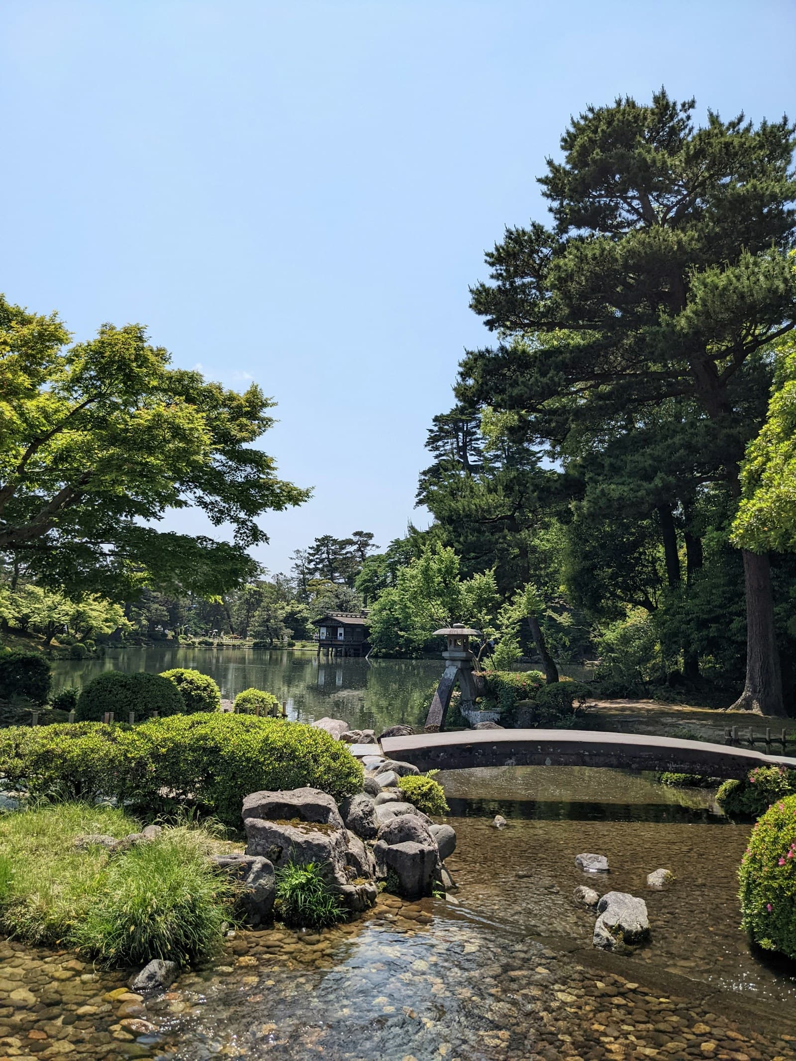 A stone bridge spans a pond at Kenrokuen Garden, with pruned pines and trees reflected in the water.
