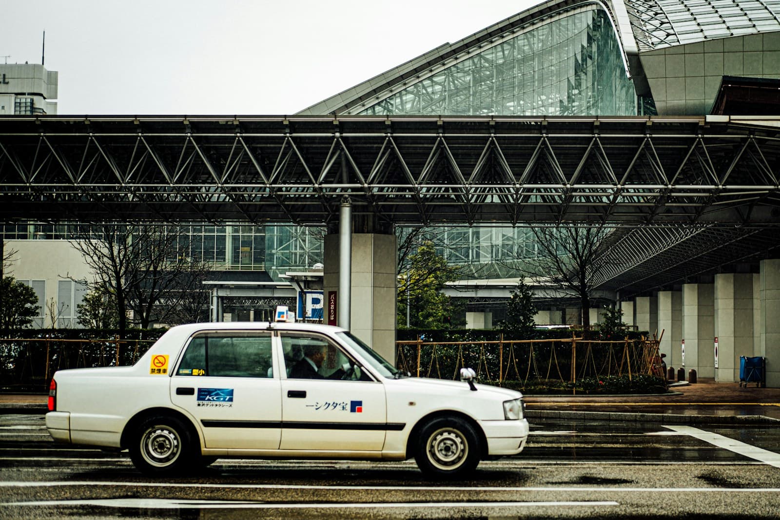 A wet urban street in Kanazawa with a white taxi and commercial buildings — ordinary city rain-day rhythm.