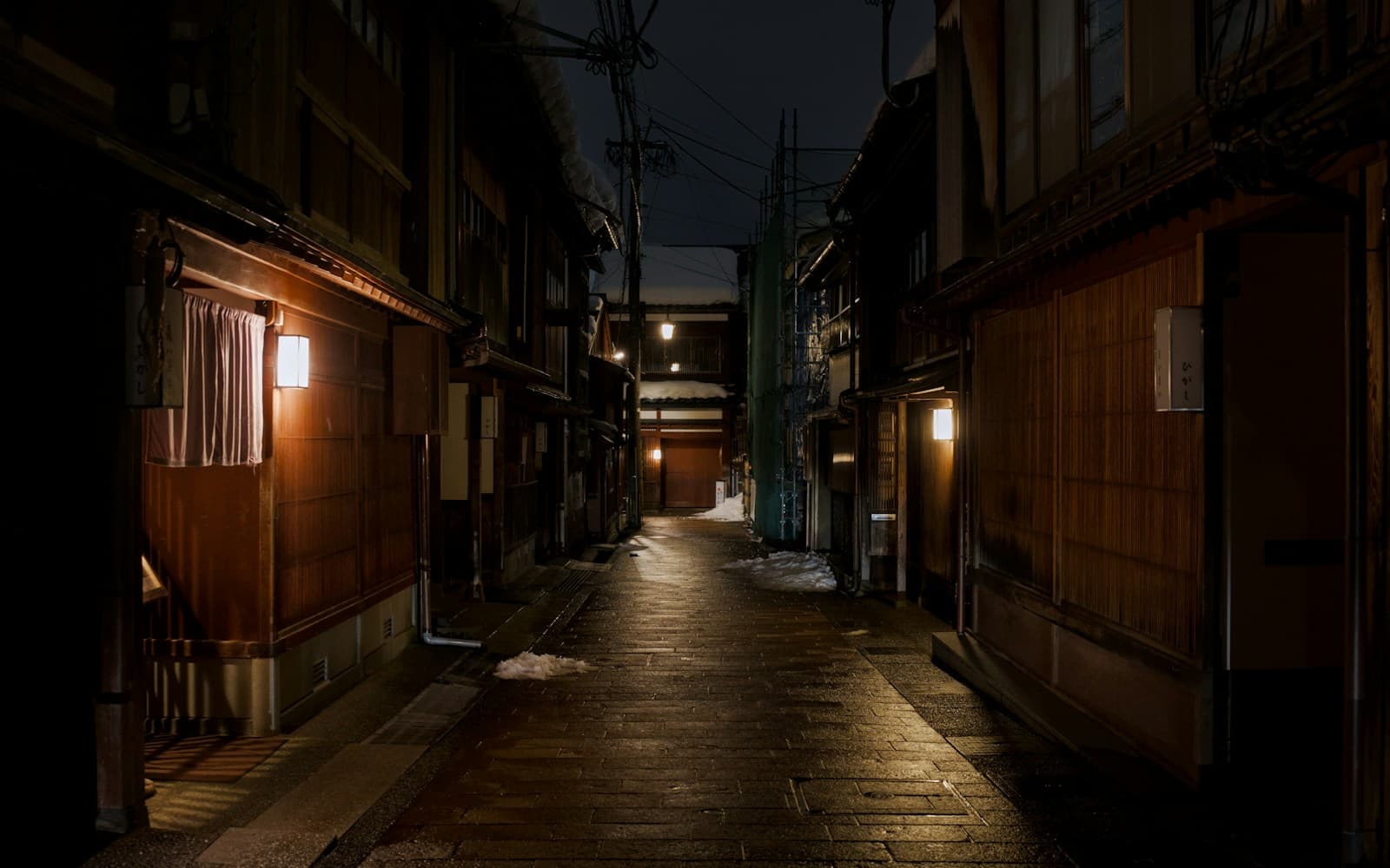 A narrow evening lane in Kanazawa's Higashi Chaya district — wooden lattice facades and warm shopfront light in a lived-in city street, not an empty postcard view.