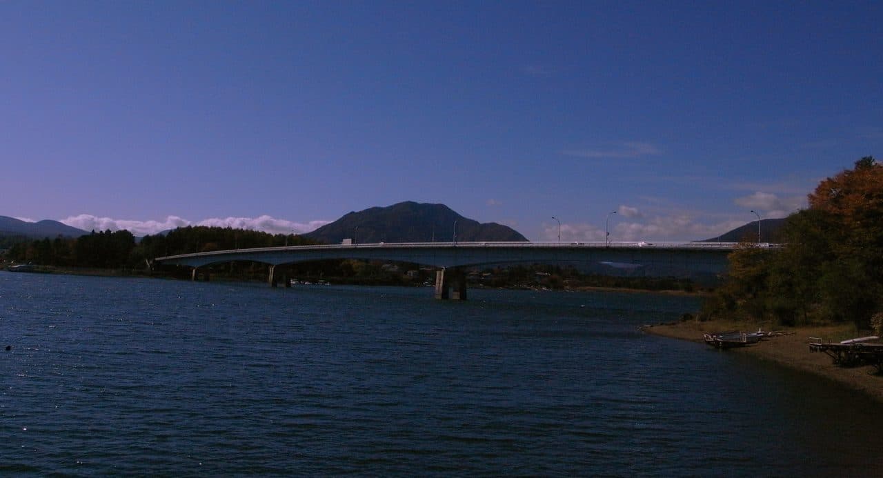 The Kawaguchiko Ohashi Bridge spans the lake, with a small shoreline beach and moored boats in the foreground and autumn trees on the bank.