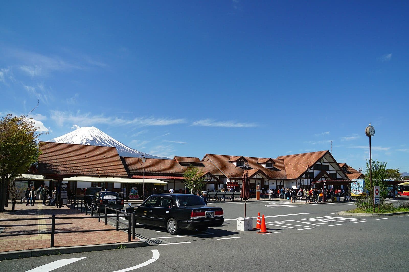Taxis and tour buses fill the forecourt of Kawaguchiko Station, with snow-capped Mount Fuji visible in the background.