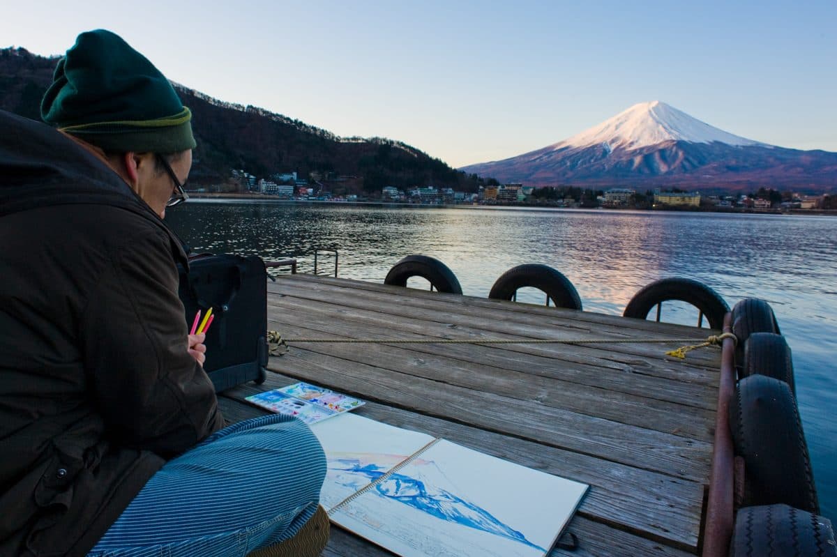 A visitor sketches Mount Fuji from a wooden pier on Lake Kawaguchiko at dusk, with the lakeside town visible along the far shore.