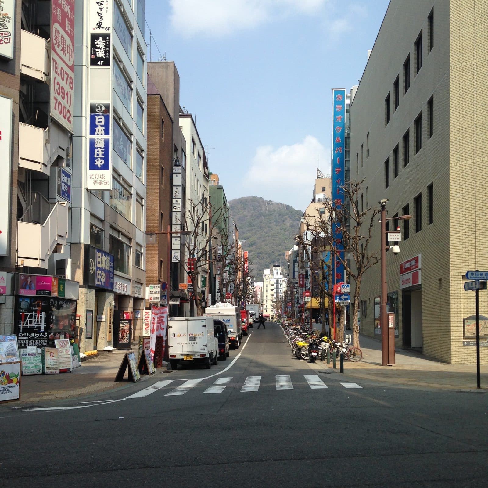 A central Kobe commercial street near Sannomiya Station, lined with shops and signage, with Mount Rokko visible at the far end.