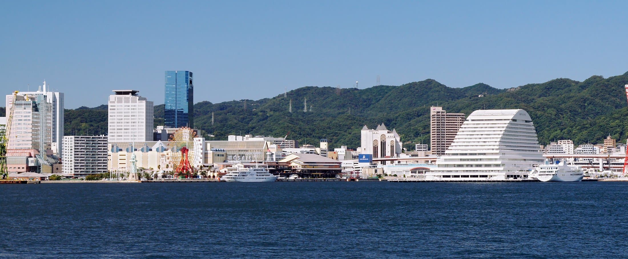 Kobe's skyline and port seen across the harbor, with Kobe Port Tower and city buildings set against the forested Rokko mountain range behind.