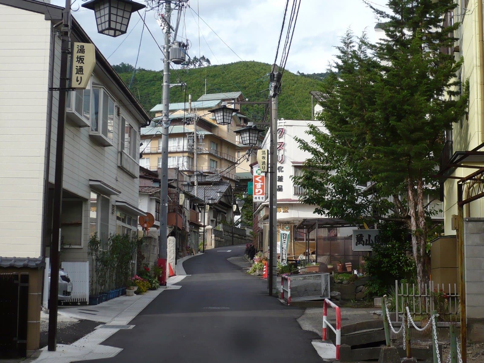 A quiet sloped street in Asama Onsen lined with ryokan buildings and lanterns, with a wooded hillside rising behind.