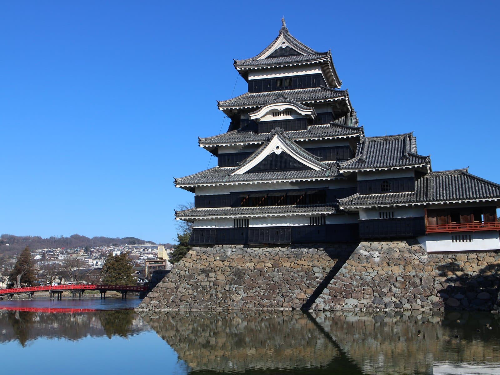 Matsumoto Castle's black keep tower rises above its stone-walled moat, with a red bridge crossing the water in the foreground under a clear blue sky.