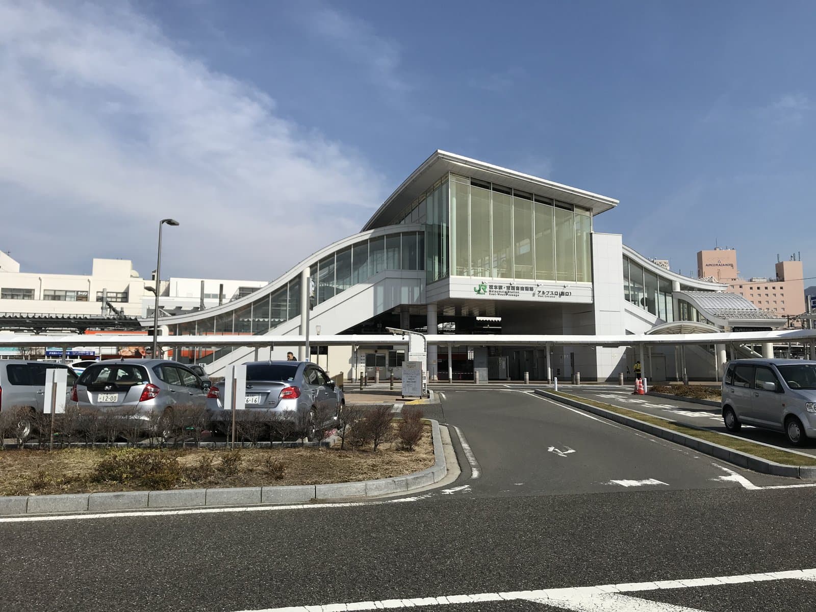 The modern glass-fronted west entrance of Matsumoto Station on a clear day, with its curved canopy and station plaza.