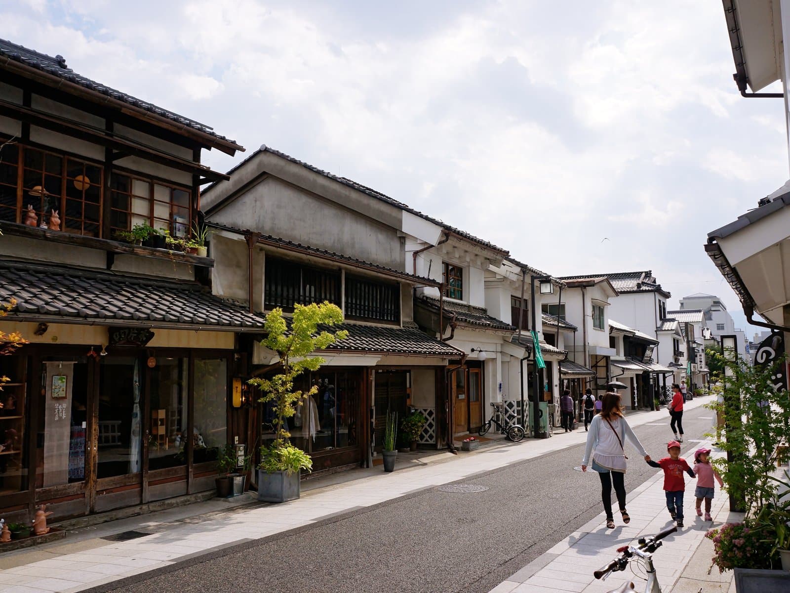 A row of white-plastered kura-zukuri storehouse buildings with tiled roofs along Nakamachi street in Matsumoto, under a partly cloudy sky.