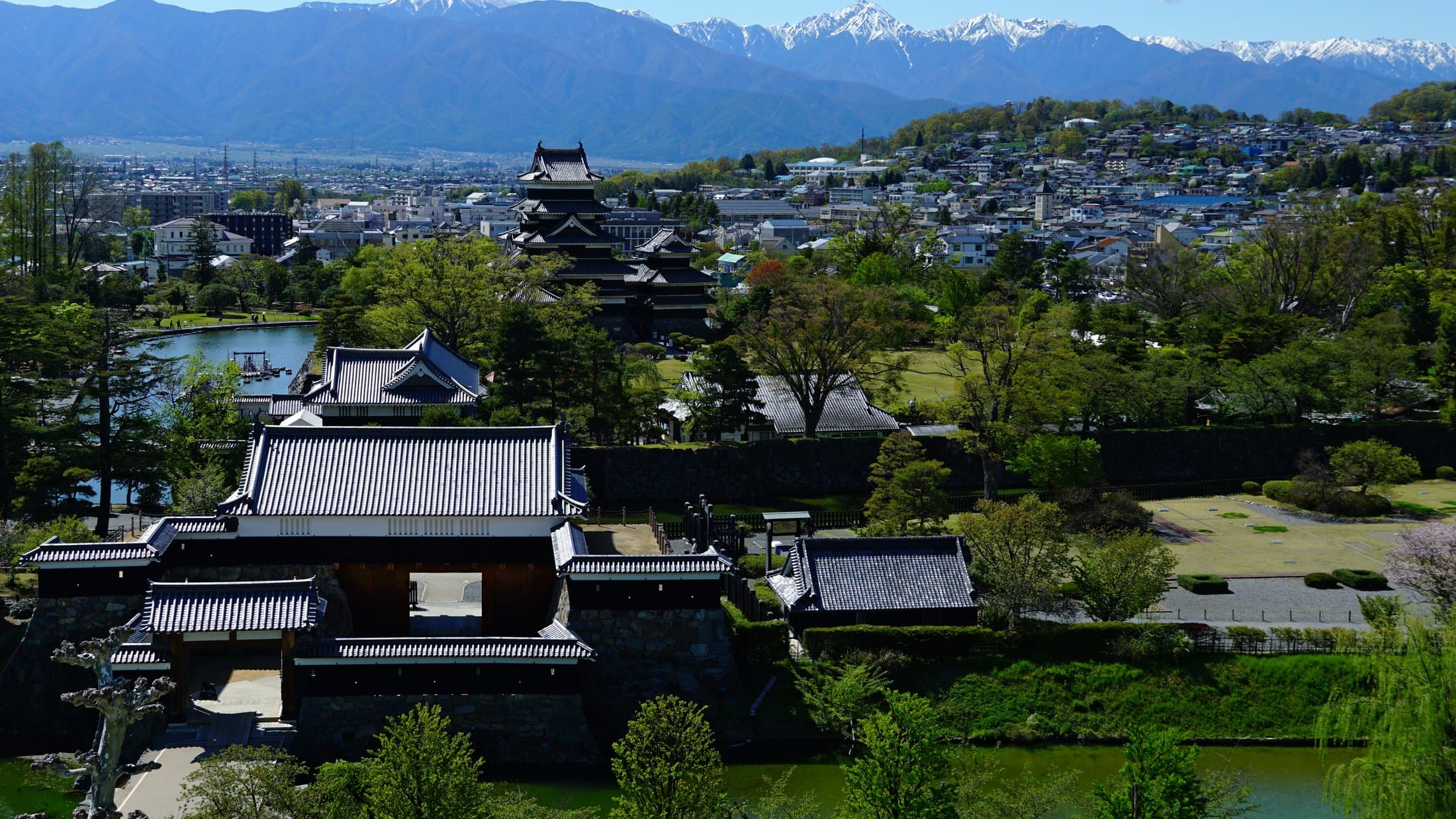 Matsumoto Castle's dark keep tower rises above the city with the snow-capped Japan Alps visible behind it under a clear sky.